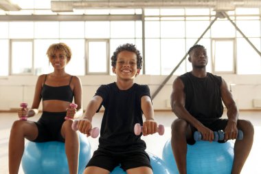 Pilates eğitiminde Afro-Amerikalı genç aile dambılları kaldırıyor ve fitness topuna oturuyor, çocuk ailesiyle yoga yapıyor.