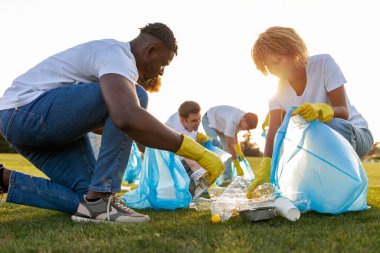 Çok ırklı bir grup gönüllü çöp torbalarıyla parkta çöp ve plastik topluyor, Afro-Amerikan erkek ve kadın şişe ve yiyecek ambalajlarını kaldırıyor, yeşil gezegen