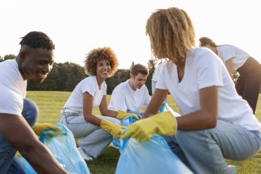 Çöp torbalarıyla çok ırklı bir grup gönüllü parkta çöp ve plastik topluyor, öğrencilerden oluşan bir grup temiz çevre, yeşil gezegen kavramı ve plastik yasağı