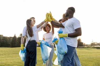 Eldiven giymiş gönüllüler ve çöp torbalarıyla birlikte başarının sevinci ve beşlik çakışmasıyla, bir grup çok ırklı insan el ele tutuşup çöpten ve plastikten parkı temizliyor.