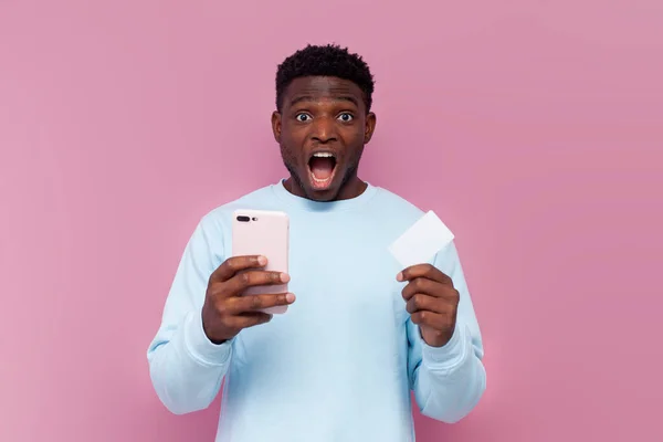 shocked african american man in blue sweater uses smartphone and credit card on pink isolated background and shouts, surprised man with phone pays for order online in amazement