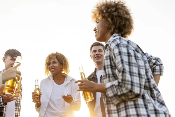 group of multiracial friends at party chatting and drinking beer outdoors, african american women and men talking and laughing and holding bottle, group of people having fun