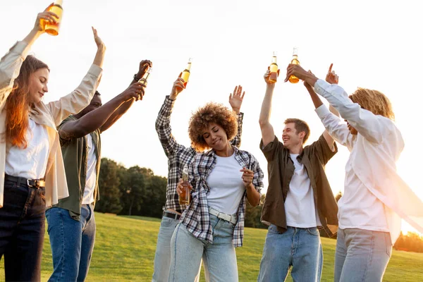 group of young multiracial students holding beer bottles and dancing at outdoor party, interracial people relaxing singing and having fun to music in park at festival with arms raised