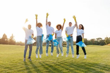 Çok ırklı bir grup gönüllü, çöp torbalarıyla eldiven giyip parkta çöp ve plastik topluyorlar. Zıplayan ve el sallayan öğrencilerden oluşan bir ekip. Gençler çevreye önem veriyor.