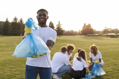 Eldivenli Afro-Amerikalı bir adam çöp torbası tutuyor ve gülümsüyor. Gönüllüler çöpten, plastikten ve çevresel bakım konseptinden parkı temizliyor.