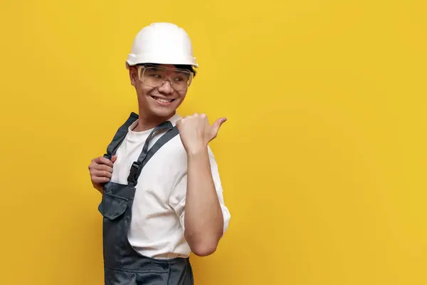 asian male construction worker in uniform and safety glasses pointing back on yellow isolated background, korean repairman in hard hat and overalls showing and advertising copy space