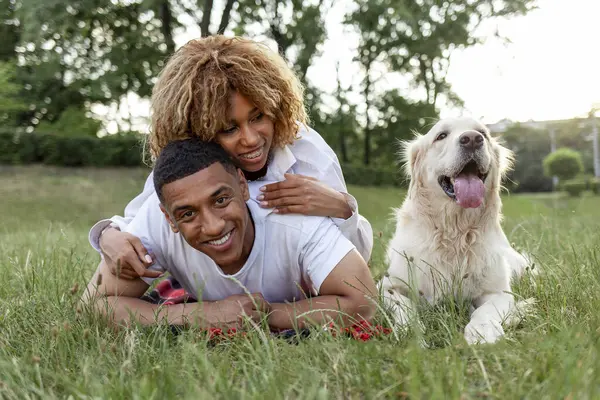 african american guy with girl lie together with golden retriever in the park in summer, young couple walks and plays with dog in nature, happy family with pet