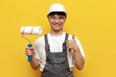 Üniformalı Asyalı ressam silindir tutuyor ve sarı izole edilmiş arka planda Koreli erkek müteahhit ve tulumlu tamirci aletleri gösteriyor.