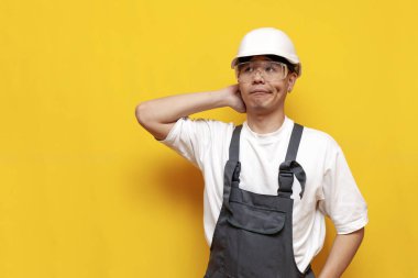 Üniformalı ve güvenlik gözlüklü kafası karışmış Asyalı adam sarı yalıtılmış arka planda düşünür ve hatırlar.