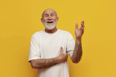 Beyaz tişörtlü yaşlı kel büyükbaba merhaba diyor ve sarı izole arka planda elini kaldırıyor, yaşlı emekli hoş bir jest yapıyor ve çağrılar yapıyor.