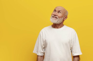 Beyaz tişörtlü yaşlı ve kel büyükbaba sarı izole bir arka plan düşünüyor ve planlıyor, düşünceli yaşlı emekliler hayal ediyor ve hayal kuruyor ve yukarı bakıyor.