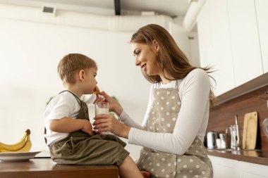 Anne önlüklü çocuk besliyor, genç kadın aşçı küçük oğluna kurabiye ve süt veriyor ve sabah evde yiyor, çocuk ailesiyle kahvaltı ediyor.