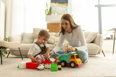 Genç bir anne ve küçük oğlu evde halıya oturur ve odada oyuncaklarla oynarlar, çocuklu kadın öğretmen yerde bir sürü oyuncak gösterir.