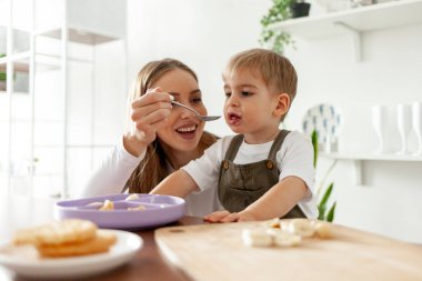 Genç anne küçük oğlunu mutfakta besliyor, kadın çocuğa ve çocuğa kaşıktan kahvaltı hazırlıyor ve gülümsüyor.