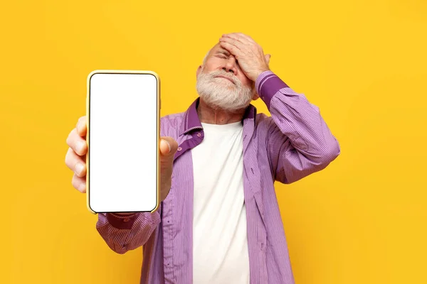 Confused old bald grandfather shows facepalm and blank smartphone screen on yellow isolated background, pensioner with phone shows gesture of failure and mistake