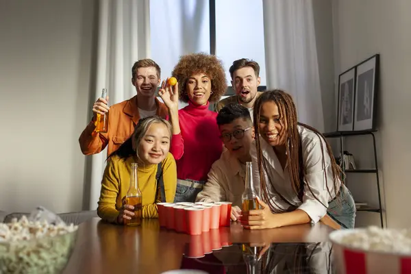 multiracial group of young student friends playing beer pong together and drinking beer at party at home, group of young people rejoicing and having fun in the room and celebrating the weekend