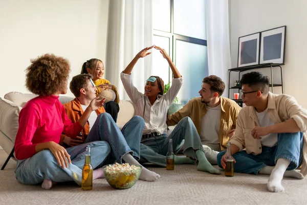 multiracial group of young student friends playing charades together and drinking beer at party at home, youth sitting on the floor with popcorn and guessing the character from the game