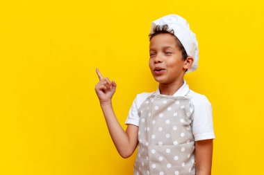 Afro-Amerikalı şef üniformalı ve şapkalı çocuk ellerini yana uzatmış, sarı izole edilmiş arka planda gülümsüyor, önlüklü genç çocuk önlükleri ve reklamları kopyalıyor.