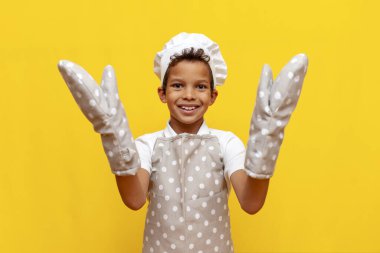 Üniformalı Afrikalı Amerikalı fırıncı ve aşçı şapkalı çocuk sarı izole edilmiş arka planda elleri boş duruyor termal eldiven giyen genç çocuk termal eldiven gösteriyor ve fotokopi alanı reklamı yapıyor.