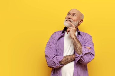 Mor gömlekli yaşlı, dalgın, kel büyükbaba kollarını sarı izole arka planda çapraz duruyor ve düşünüyor, yaşlı emeklilerin hayalleri ve planları var ve başka yere bakıyor.