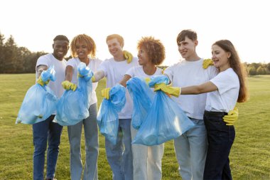 Çöp torbalarıyla eldiven giyen çok ırklı bir grup gönüllü parkta çöp ve plastik topluyor, öğrencilerden oluşan bir grup gezegene yardım ediyor, gençlik çevreyle ilgileniyor.