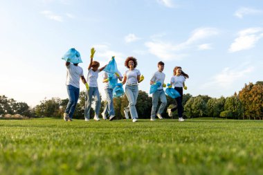 Çok ırklı gönüllüler eldiven takmış ve parkta çöp torbalarıyla çöp toplamak için koşuyor, bir grup genç plastik, çevre bakımı ve yeşil gezegeni temizlemeye gidiyor.