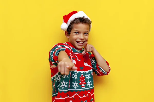I choose you. african american boy in christmas sweater and santa hat points with his hands forward on yellow isolated background, teenage child in christmas clothes points at the camera