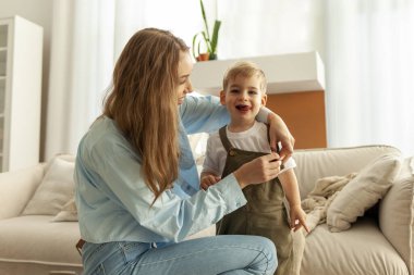 Genç anne, küçük oğlunun evde elbisesini giymesine yardım ediyor. Kadın da çocuğa bakıyor ve onu giydiriyor.