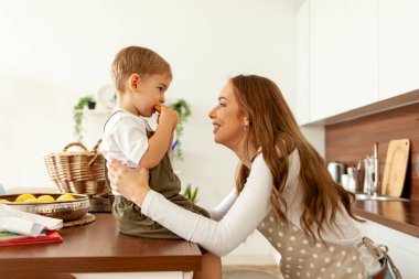 Mutfakta önlüklü genç bir anne küçük oğlunu kurabiyelerle besler, kadın çocuğuyla birlikte hamur işi yer, 2 yaşındaki çocuk ailesiyle kahvaltı eder.