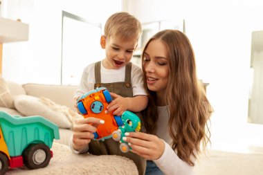 Genç anne küçük oğluyla evde oyuncaklarla oynuyor, çocuklu kadın yerde oyuncak araba kullanıyor, 2 yaşındaki çocuk ailesiyle eğleniyor.