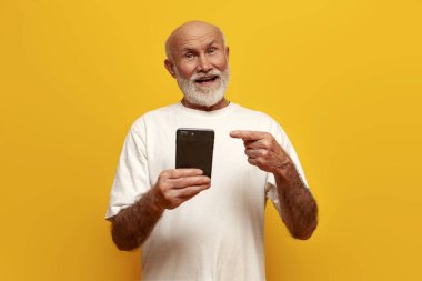 Beyaz tişörtlü yaşlı kel büyükbaba sarı izole edilmiş arka planda akıllı telefon kullanıyor. Yaşlı adam cep telefonunu işaret ediyor ve gülümsüyor.