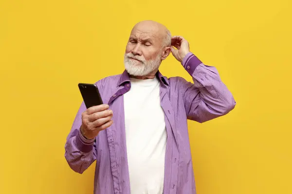 puzzled old bald grandfather in purple shirt uses smartphone on yellow isolated background, confused old pensioner looks at the phone online and scratches his head