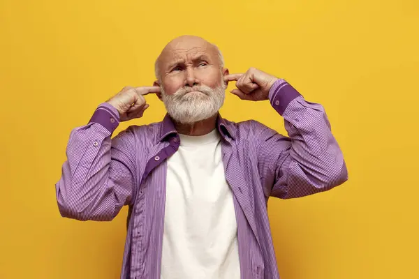 offended old bald grandfather in purple shirt covers his ears with his hands and ignores on yellow isolated background, sad old man pensioner does not listen and avoids