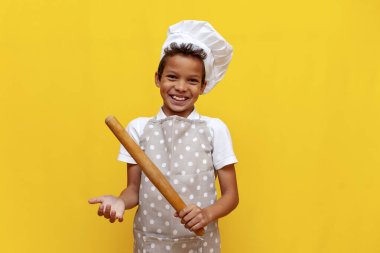 Afro-Amerikalı aşçı üniformalı, şapkalı, elinde mutfak eşyaları olan, sarı izole edilmiş arka planda gülümseyen, önlüklü, kameraya bakan genç bir çocuk.