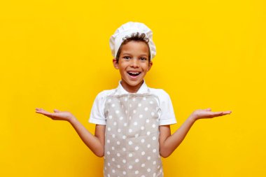 Üniformalı ve aşçı şapkalı Afro-Amerikan çocuk sarı izole bir arka plan seçiyor, önlüklü genç ellerini kaldırıyor ve kopya alanı gösteriyor.