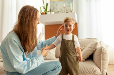 Genç anne küçük oğlunu elinden tutar ve saçlarını düzleştirir. 2 yaşında, açık elbiseli bir çocuk gülümser ve kameraya bakar. Kadın çocuğa bakar.
