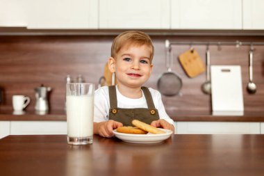 2 yaşındaki küçük çocuk mutfakta masada oturuyor ve sütle kurabiye yiyor, çocuk kahvaltısını evde yapıyor ve gülümsüyor ve kameraya bakıyor.