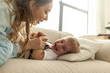 Genç anne küçük oğluyla kanepede oynuyor ve ellerini tutuyor. 2 yaşındaki çocuk gülüyor ve ailesiyle eğleniyor.