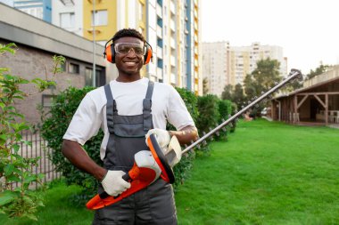 Bahçe işçisinin portresi elektrikli aletle, koruyucu gözlüklü ve kulaklıklı Afro-Amerikan adam bahçede fırça makası tutuyor ve gülümsüyor.