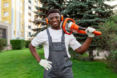 Bahçe işçisinin portresi elektrikli aletle, koruyucu gözlüklü ve kulaklıklı Afro-Amerikan adam bahçede fırça makası tutuyor ve gülümsüyor.