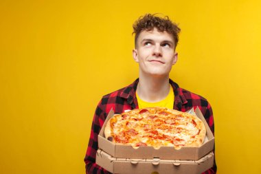 young curly cute guy with pizza looking away and dreaming, pensive man holding pizza on yellow background