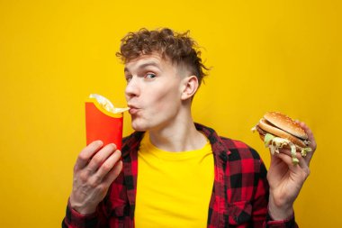 Hamburgerli genç kıvırcık adam patates kızartması yiyor ve kameraya şaşkınlıkla bakıyor, sarı arka planda fast food yiyen bir adam.