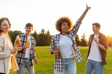 Bir grup genç çok ırklı öğrenci ellerinde bira şişeleri ve açık hava partisinde dans ediyorlar, ırklar arası insanlar şarkı söylüyor ve festivalde parkta müzik eşliğinde eğleniyorlar.