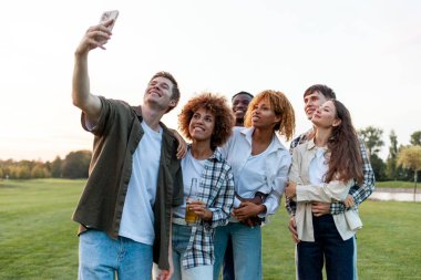 Bir grup çok ırklı genç akıllı telefondan selfie çekiyor ve parkta fotoğraf çekiyor. Irklar arası öğrenciler açık havada video iletişimiyle iletişim kuruyor.