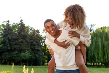 Afro-Amerikan mutlu çifti yazın parkta birlikte yürüyor ve gülümsüyor, erkek kadını sırtında taşıyor ve doğa, özgürlük ve ilişki kavramında sarılıyor.