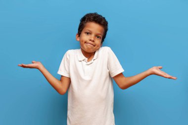 Emin olmayan kıvırcık saçlı, beyaz tişörtlü Amerikalı çocuk sarı izole edilmiş arka planda omuzlarını silkeliyor, kafası karışmış 9 yaşındaki çocuk ellerini kaldırıyor ve farkında değil.