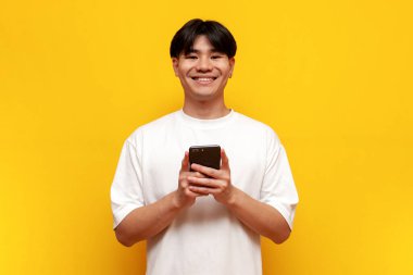 Beyaz tişörtlü genç Asyalı öğrenci sarı izole edilmiş arka planda akıllı telefon kullanıyor, Koreli adam cep telefonu tutuyor ve gülümsüyor.