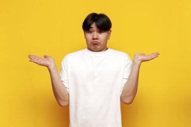 Beyaz tişörtlü Asyalı genç adam sarı yalıtılmış arka planda omuzlarını silkiyor, kafası karışmış Koreli adam bunu bilmiyor ve ellerini açıyor.