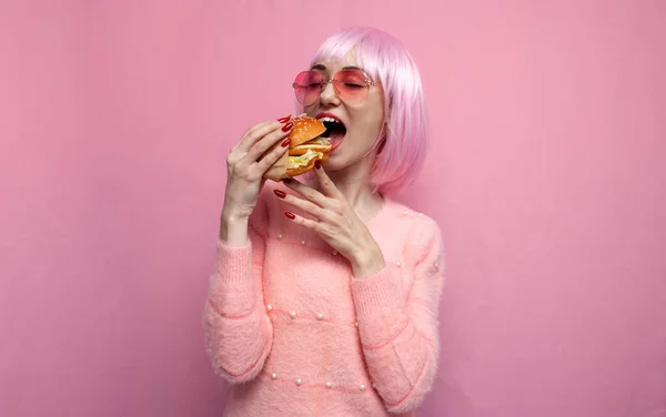 young cute girl with pink short hair eats a burger with her mouth wide open, a woman with a short haircut eats fast food on a pink background