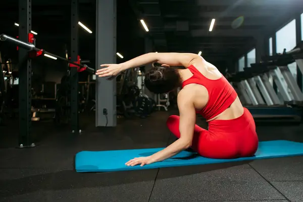 Kırmızı spor kıyafetli atletik bir kadın yoga matte 'inde oturuyor ve ısınıyor, yoga yapıyor ve esniyor, fitness kulübünde esnek egzersiz yapan çekici bir kadın.